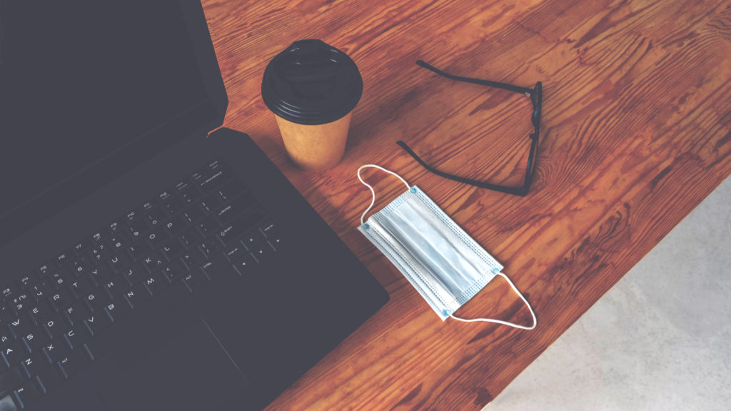 Desk with a laptop, coffee cup, glasses and medical mask on it.
